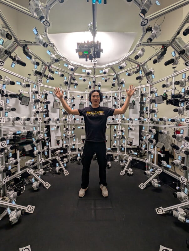 A person stands with arms raised inside a circular room filled with numerous cameras and lights mounted on metal frames, likely for 3D scanning or motion capture. The person wears a black "Pennzoil" t-shirt and glasses.