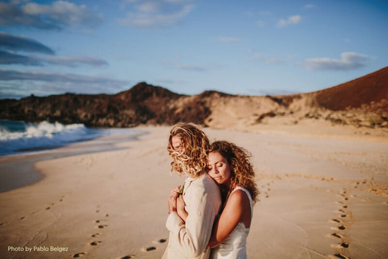 A couple embraces on a sandy beach with rocky hills in the background. The woman, in a white dress, hugs the man from behind as they both stand in footprints along the shore under a partly cloudy sky.