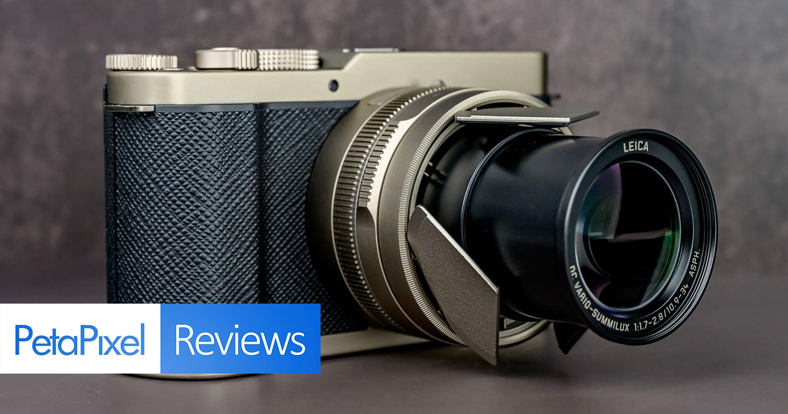 A Leica camera with a textured grip and extended zoom lens sits on a dark surface. The image features a "PetaPixel Reviews" banner in the lower left corner.