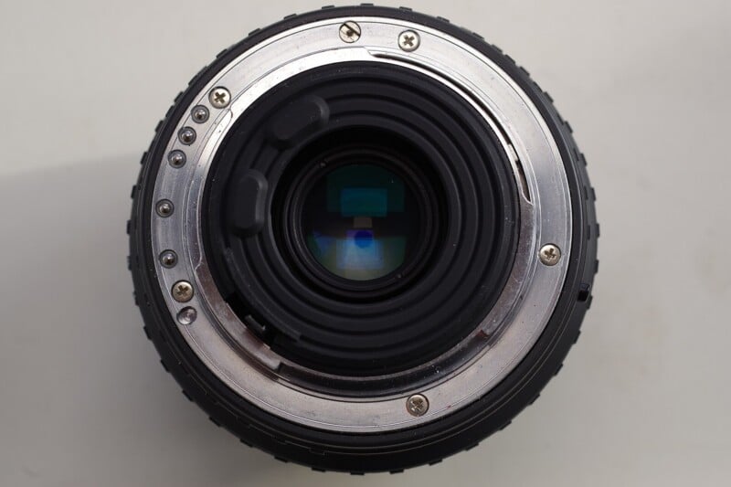 Close-up view of the rear side of a camera lens showing the metal lens mount, glass elements, and mounting screws against a plain light background.