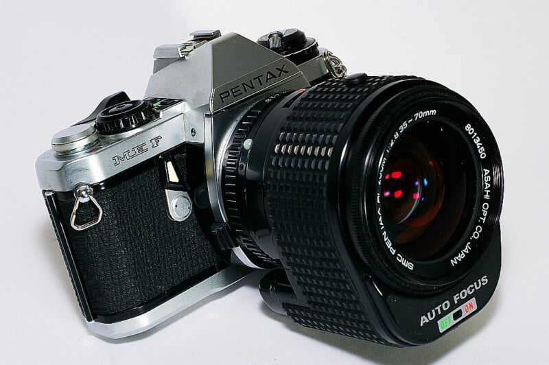 A close-up photo of a vintage Pentax ME F film camera with a large, black autofocus lens attached, resting on a white surface.