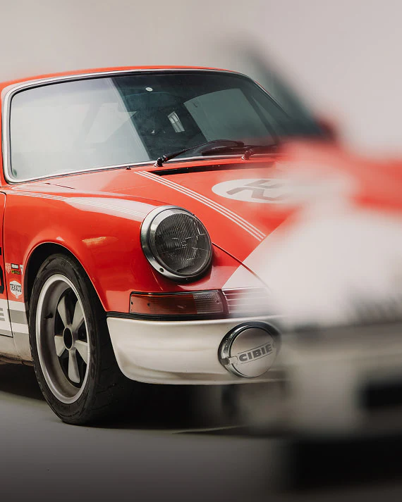 Close-up of a vintage red sports car with a round headlight and black wheels, partially obscured by a blurred white object in the foreground. Racing decals and a Cibie fog light are visible on the car.