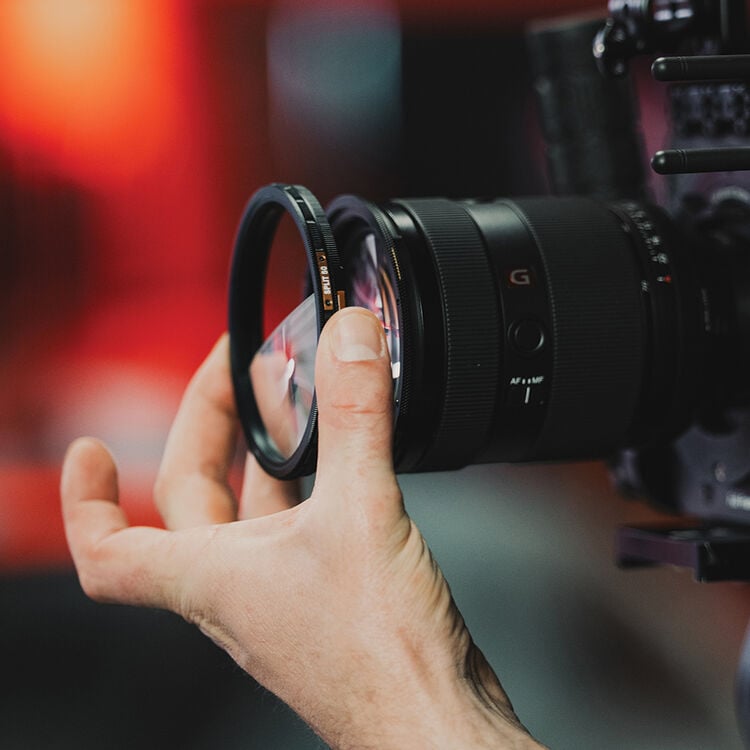 A hand holds a camera lens filter in front of a black camera lens, preparing to attach it. The background is blurred with warm red tones.