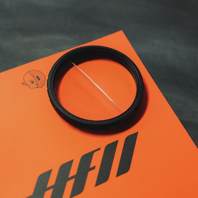 A round camera filter with a visible crack lies on an orange surface with black diagonal stripes and a small cartoon face in the corner.
