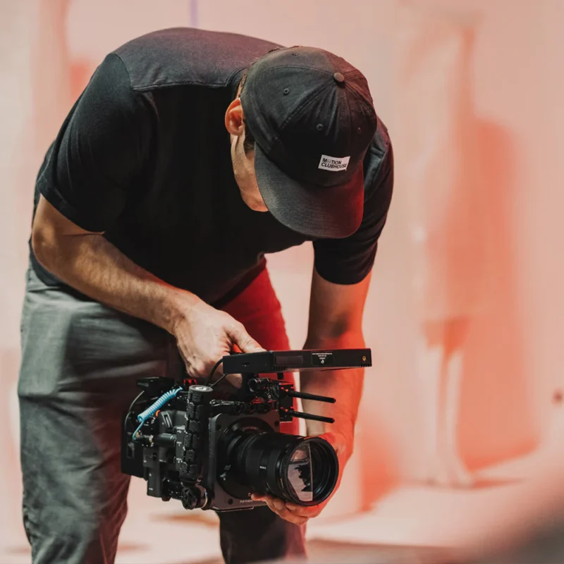 A person wearing a black cap and t-shirt holds and adjusts a professional video camera, aiming it downward in a softly lit, reddish room.