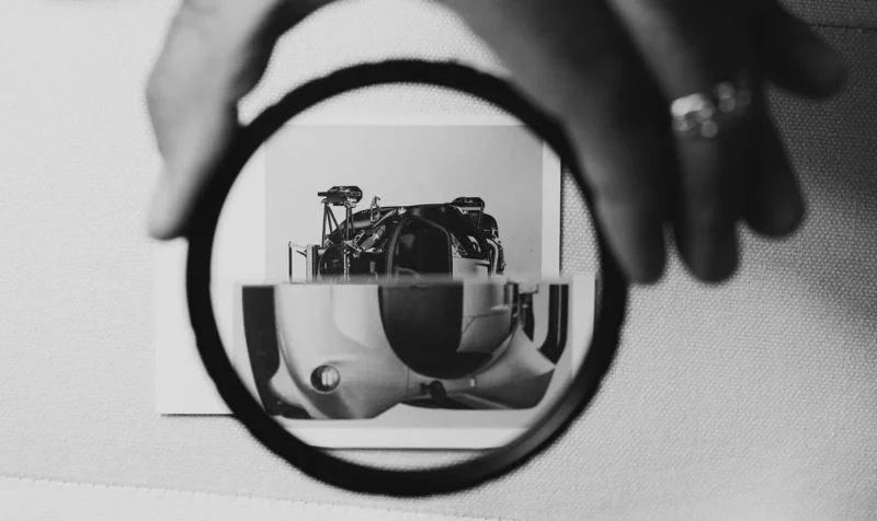 A hand holds a round magnifying glass over a black and white photograph of a car, enlarging and distorting the lower half of the image.
