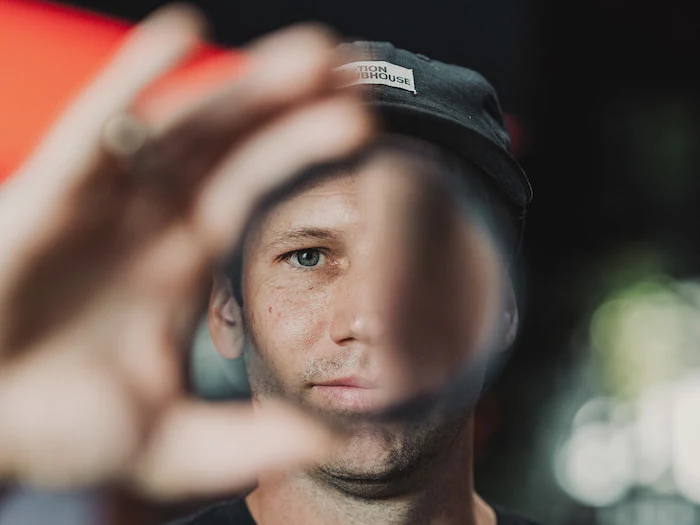 A person wearing a black cap holds a round glass filter in front of their face, partially distorting their facial features through the lens. The background is blurred.