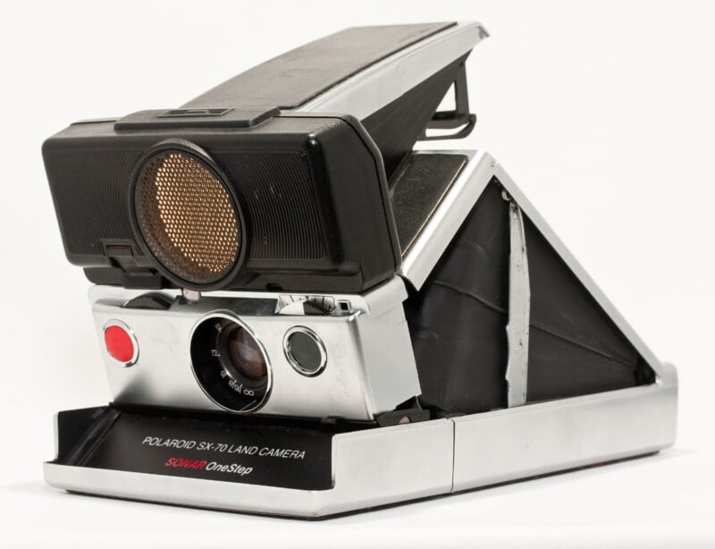 A vintage Polaroid SX-70 Land Camera in silver and black, with its lens and viewfinder visible and the camera in its open, ready-to-use position against a white background.
