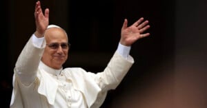 A man dressed in white religious attire, including a skullcap and glasses, stands with both arms raised, smiling and appearing to greet or bless a crowd.