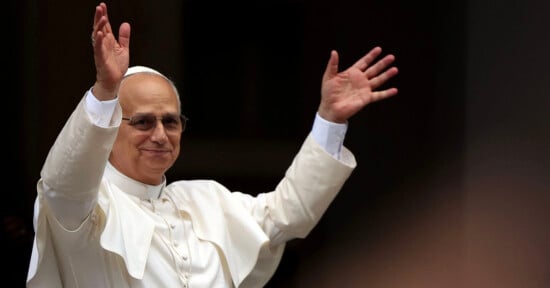 A man dressed in white religious attire, including a skullcap and glasses, stands with both arms raised, smiling and appearing to greet or bless a crowd.