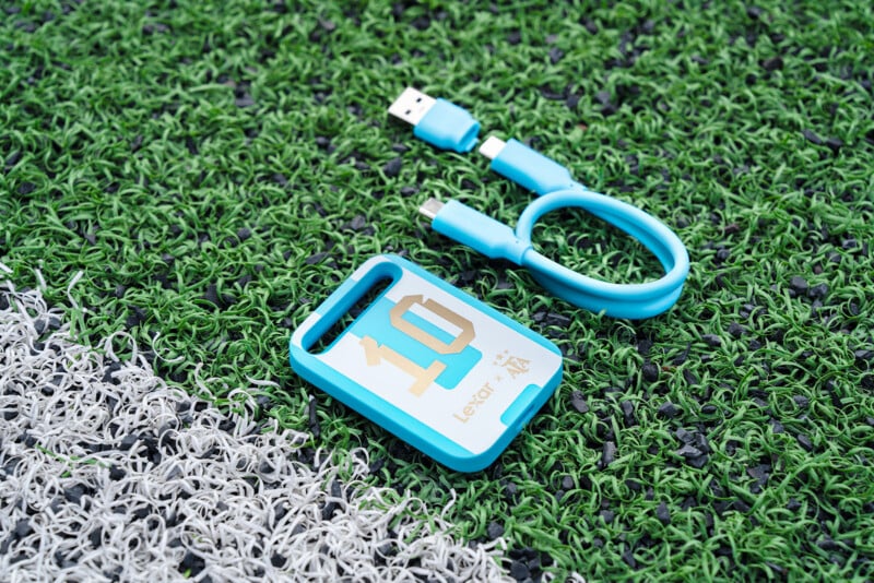 A blue USB Type-C card reader and a short blue USB cable rest on artificial grass near a white boundary line.