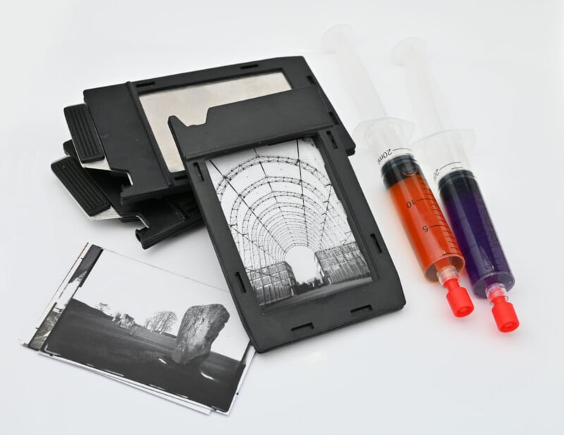 A stack of black photographic film holders, black-and-white photo prints, and two syringes filled with red and purple liquids are arranged on a white surface.