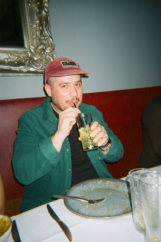 A person in a red cap and green shirt sits at a dining table, sipping a drink through a straw. The table has empty plates and glasses, and the person looks toward the camera. A large mirror hangs on the wall behind them.