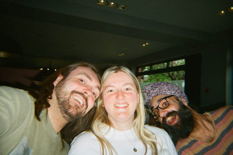 Three people smile and pose closely together indoors, with a man with long hair and a beard on the left, a woman with blonde hair in the center, and a man with glasses and a patterned headwrap on the right.
