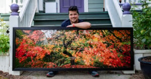 A smiling man kneels on a porch, holding a large framed photograph of a tree with vibrant red and orange autumn leaves. The house behind him has green steps, white railings, and purple accents.
