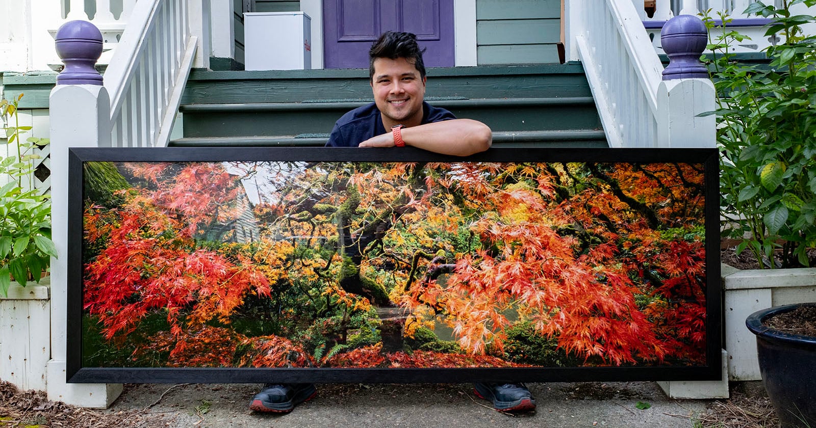 A smiling man kneels on a porch, holding a large framed photograph of a tree with vibrant red and orange autumn leaves. The house behind him has green steps, white railings, and purple accents.