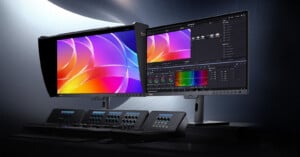 Two computer monitors display colorful graphics and editing software. Both screens are mounted on stands above an advanced video editing control panel with multiple buttons and dials.