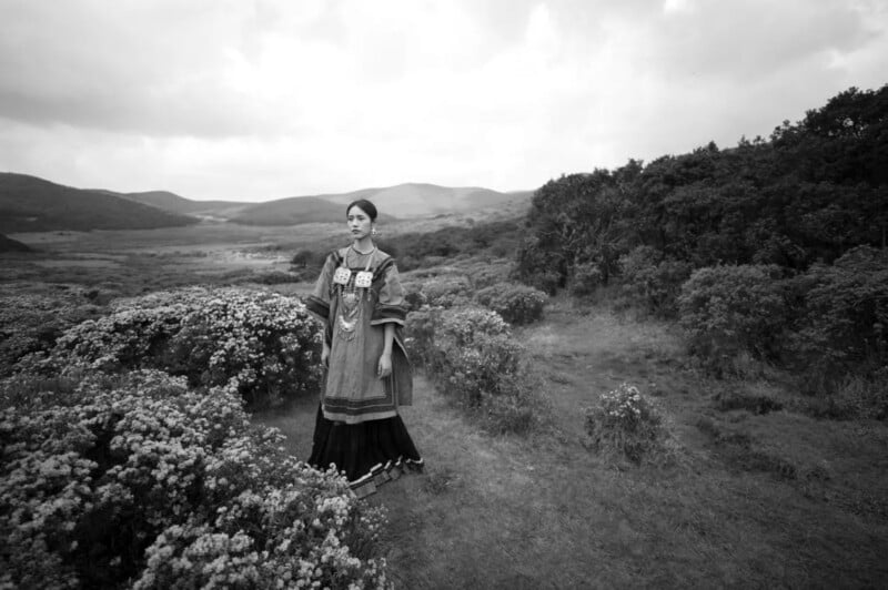 A woman in traditional clothing stands among flowering bushes in a wide, open landscape with hills in the distance under a cloudy sky. The image is in black and white.