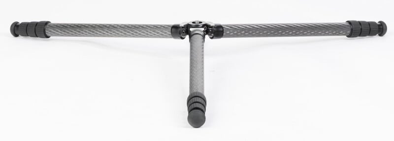 A carbon fiber tripod center column with three adjustable legs extended outward, two horizontally and one vertically, forming a T-shape on a white background.