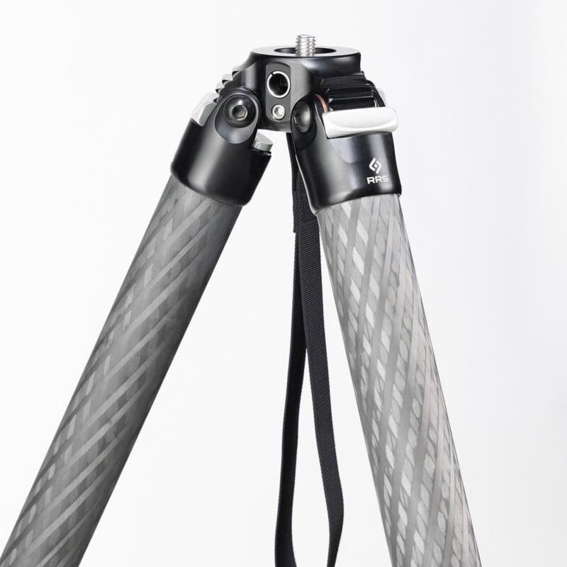 A close-up view of a carbon fiber tripod with two extended legs, metal joints, a mounting screw on top, and a black carrying strap attached to one leg.