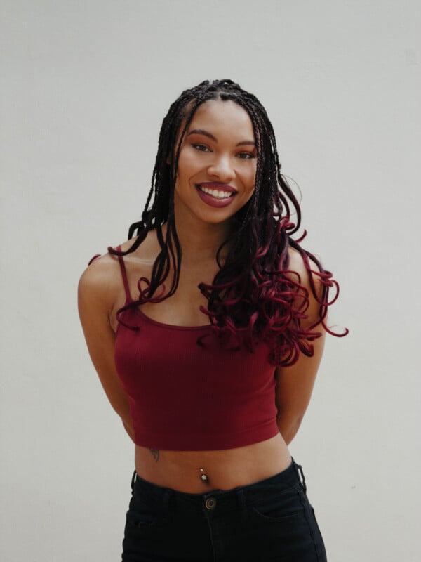 A smiling woman with long braided hair, wearing a burgundy crop top and black pants, stands against a plain white background with her hands behind her back.