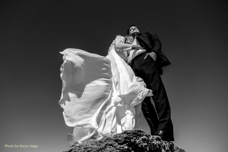 A bride and groom stand on a rocky surface against a clear sky. The bride's dress flows dramatically in the wind as they embrace, looking upwards. The photo is in black and white.