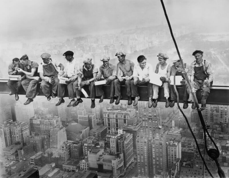 Eleven construction workers eat lunch and talk while sitting on a steel beam high above New York City, with skyscrapers visible far below and a hazy skyline in the background.