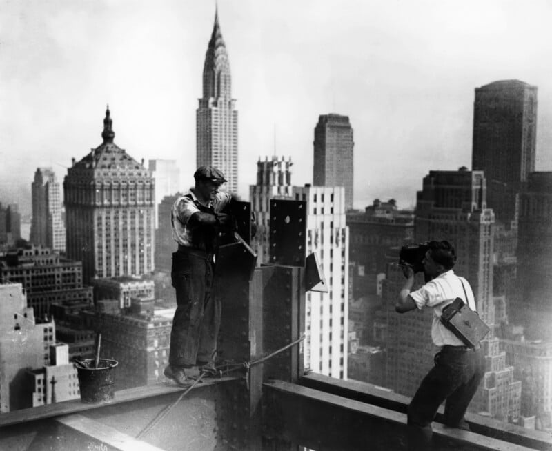 A construction worker stands on a steel beam high above New York City while a photographer takes his picture. Skyscrapers, including the Chrysler Building, fill the background under a hazy sky.
