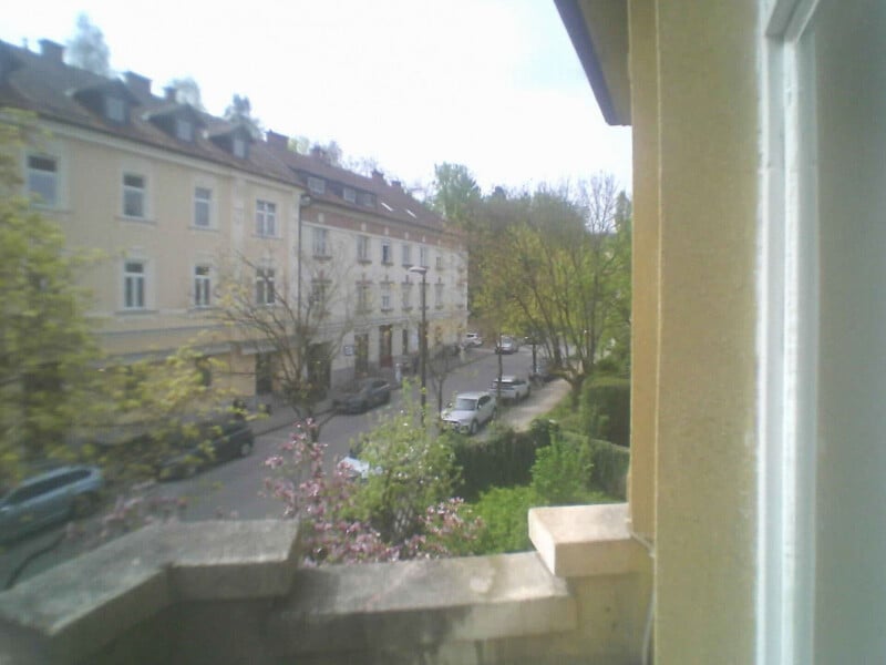 View from a balcony overlooking a quiet street lined with parked cars, trees, and light-colored residential buildings on a cloudy day.