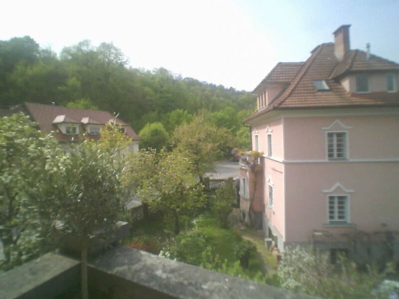 A slightly blurry view of a pink house with a red-tiled roof, surrounded by greenery and trees, with another house visible in the background under a bright sky.