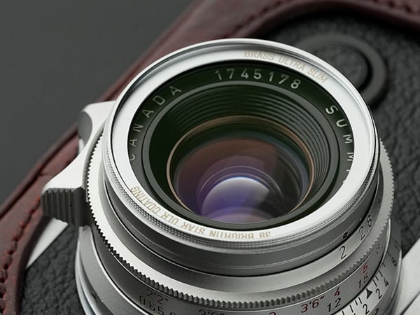 Close-up of a camera lens mounted on a black and brown leather camera body, showing reflections in the glass and engraved details including "CANADA," "SUMMITAR," and various numbers around the lens.