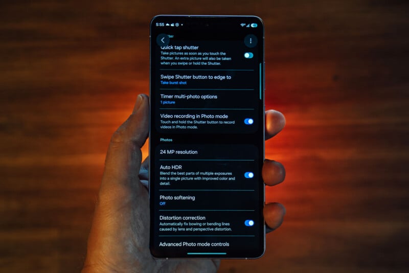 A hand holds a smartphone displaying camera settings on the screen, including options like quick tap shutter, video recording, photo resolution, HDR, photo softening, and distortion correction. The background is blurred and warmly lit.