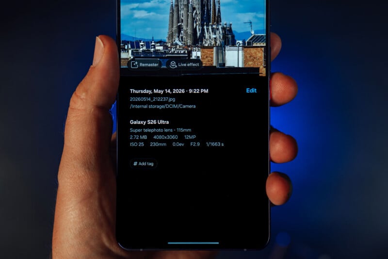 A hand holds a smartphone displaying photo details, including date, time, file name, camera model (Galaxy S26 Ultra), and camera settings; part of a cathedral is visible on the phone screen.