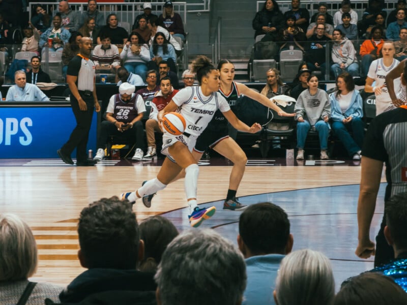 A basketball player in a white uniform dribbles the ball while being closely guarded by a defender in a dark uniform during a game, with referees and spectators watching from the stands.