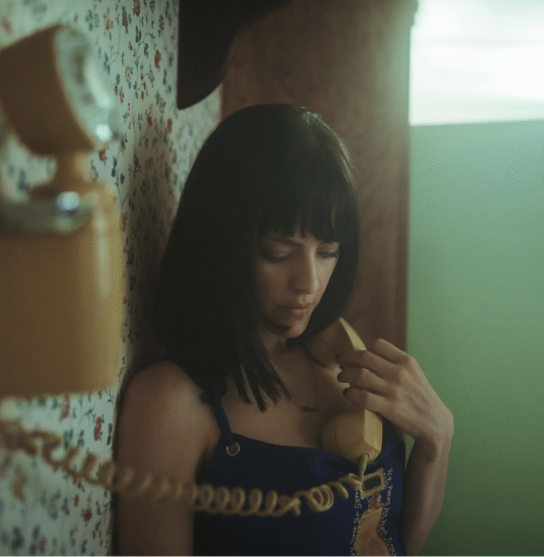 A woman with dark hair stands against a floral wallpapered wall, looking down while holding a yellow vintage telephone with a coiled cord. The lighting is soft and nostalgic.