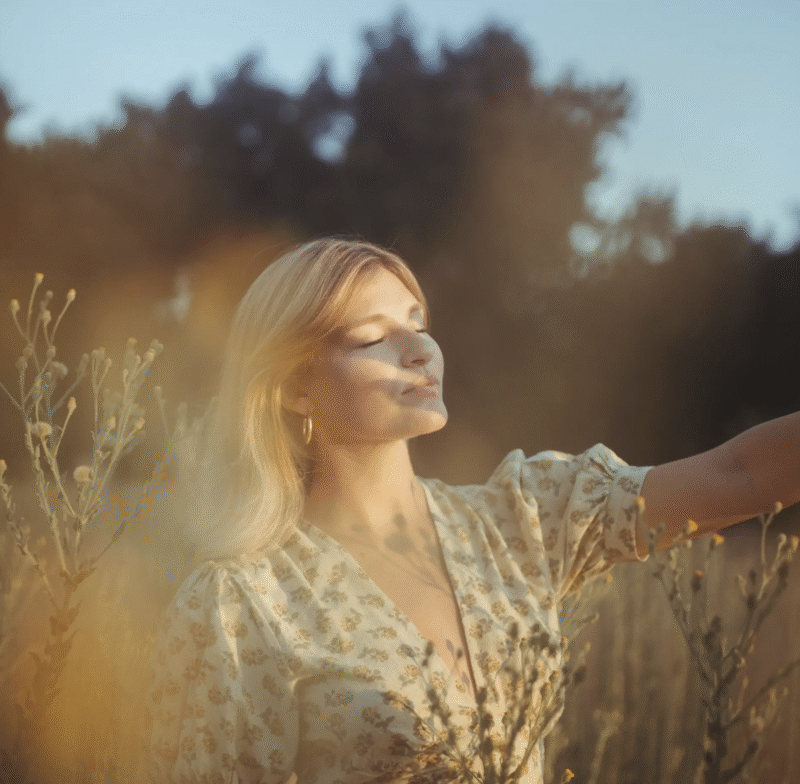 A woman with blonde hair stands in a sunlit field, wearing a light floral dress. She closes her eyes and gently smiles, with her arm outstretched among tall wildflowers. Soft golden light surrounds her.