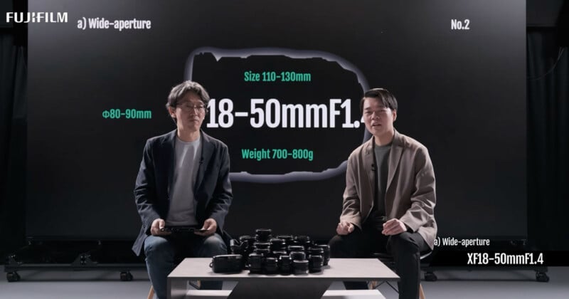 Two men sit in front of a large screen displaying camera lens specifications (18–50mm F1, size 110–130mm, weight 700–800g) with several camera lenses on a table between them. The screen shows the Fujifilm logo.