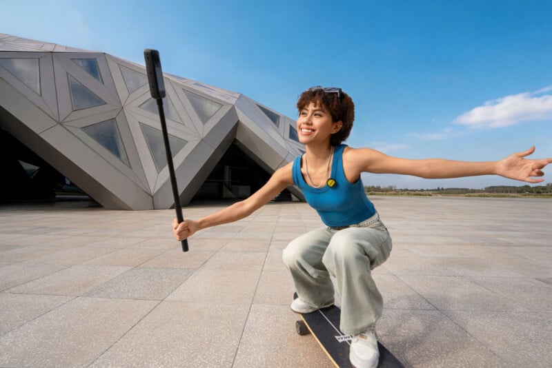 A young woman in a blue tank top and jeans skateboards on a wide, tiled plaza, holding a selfie stick and smiling, with a modern, angular building and a clear blue sky in the background.