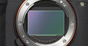 Close-up view of a digital camera's exposed sensor and metal lens mount, showing electronic contacts and screws, with part of the camera body visible on the left and a number "9" on the top right.