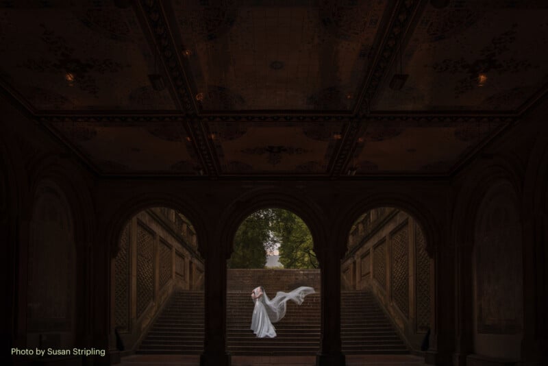 A bride in a flowing white dress stands under a grand, ornate archway with stairs and green trees behind her. Her veil is dramatically lifted, capturing a sense of motion and elegance. Warm lights illuminate the arched ceiling.
