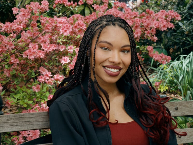 A woman with braided hair and a burgundy top smiles while sitting on a wooden bench in front of vibrant pink flowering bushes in a garden.