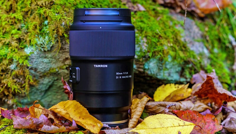 A Tamron 90mm f/2.8 macro camera lens stands upright on the ground surrounded by colorful autumn leaves and moss-covered rocks.