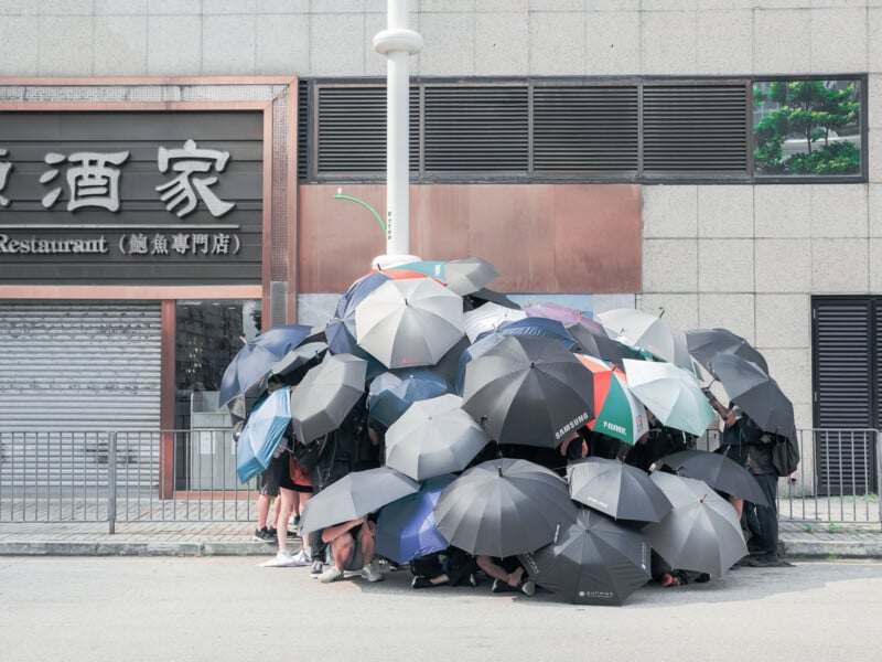 A large group of people huddles together on a sidewalk, mostly concealed under overlapping black umbrellas, in front of a building with a restaurant sign. Only their legs and shoes are visible beneath the umbrellas.