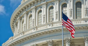 A close-up view of the United States Capitol building’s ornate facade with arched windows, and an American flag waving in the foreground against a blue sky.