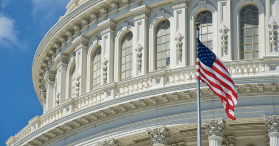 A close-up view of the United States Capitol building’s ornate facade with arched windows, and an American flag waving in the foreground against a blue sky.