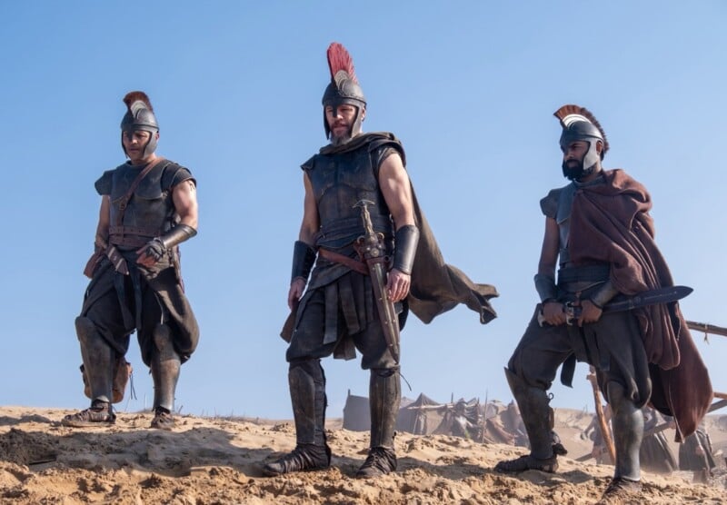 Three men in ancient warrior armor and helmets stand on sandy ground under a clear blue sky, with tents and weapons visible in the background, suggesting a historical or battle setting.
