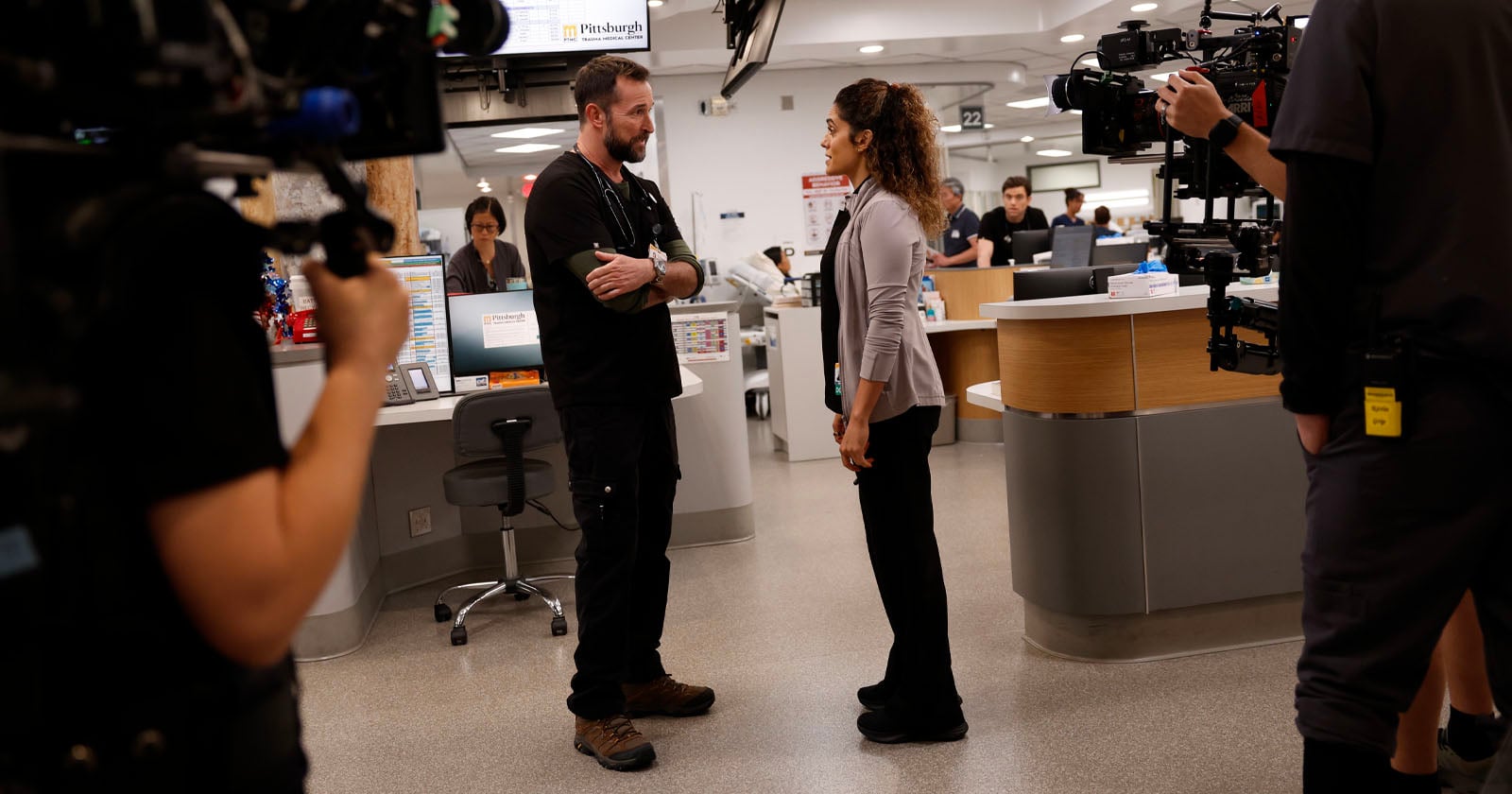 Two people stand facing each other and talking in a busy hospital setting, surrounded by medical staff and camera crew filming the scene with professional equipment.