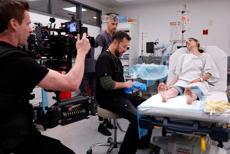 A film crew records a scene in a hospital set, showing a woman in a hospital gown lying on a bed as two men in medical attire attend to her, while a cameraman films the action up close.