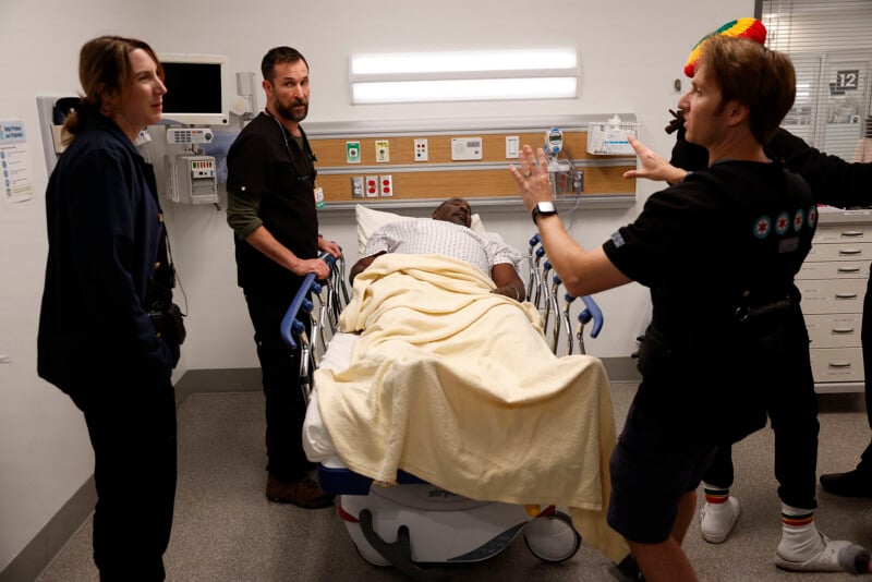 A patient lies in a hospital bed covered with a blanket, surrounded by three medical staff who appear to be discussing his care in a well-lit hospital room.