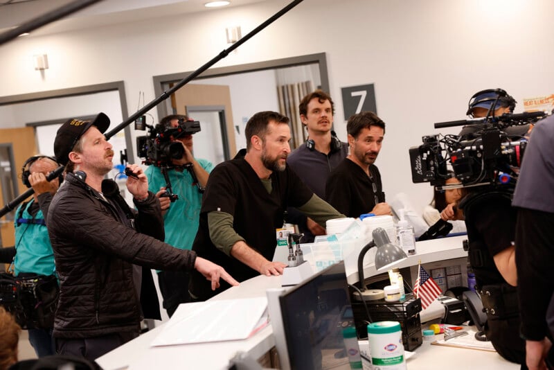 A film crew and actors gather around a hospital reception desk set, with cameras, sound equipment, and crew members actively engaged in filming a scene.